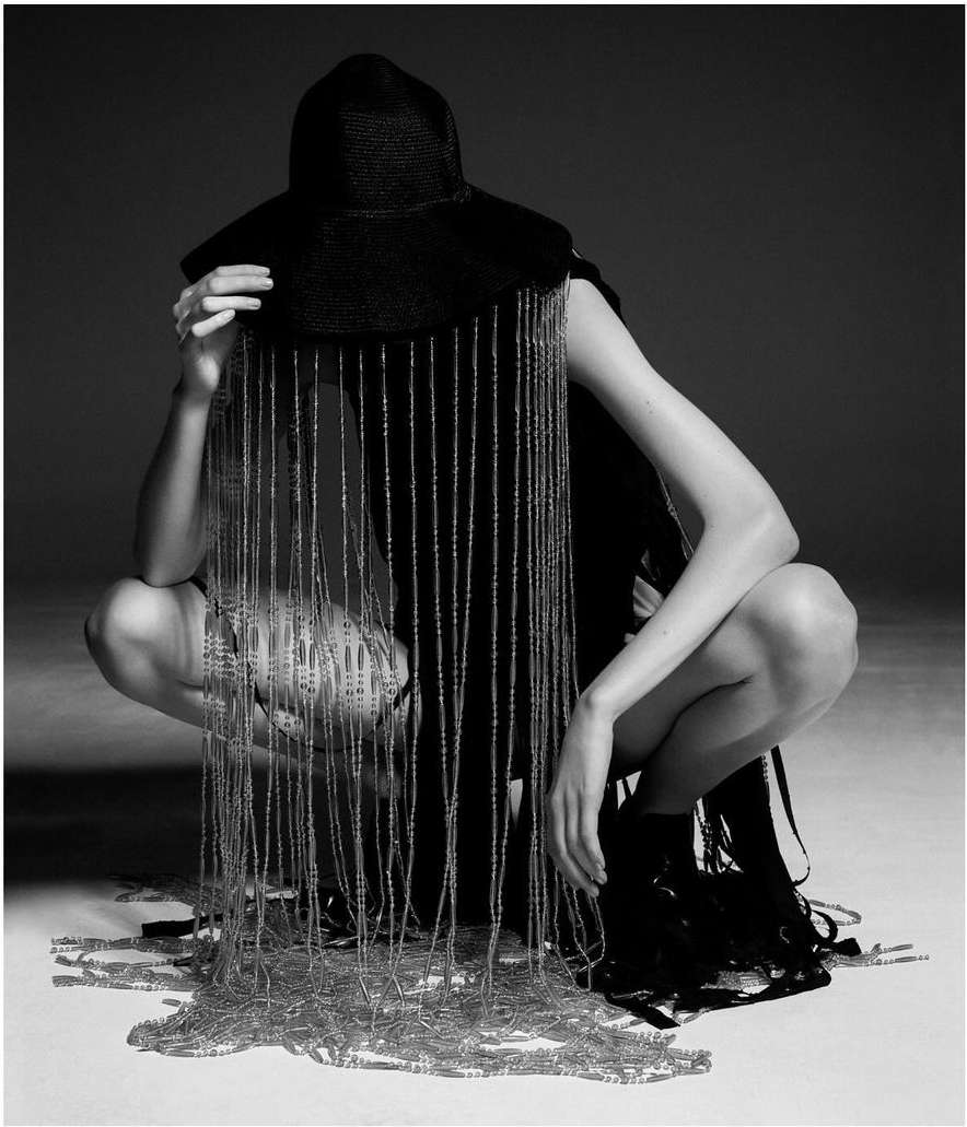 Monochrome fashion shot of a crouched model wearing a wide-brimmed hat with cascading beaded fringe, and elegant textural clothing.