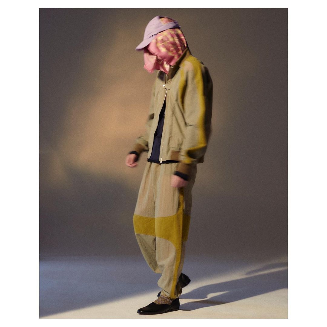 Person in beige jacket and pants with a purple hat stands in front of a neutral background in a fashion studio setting.
