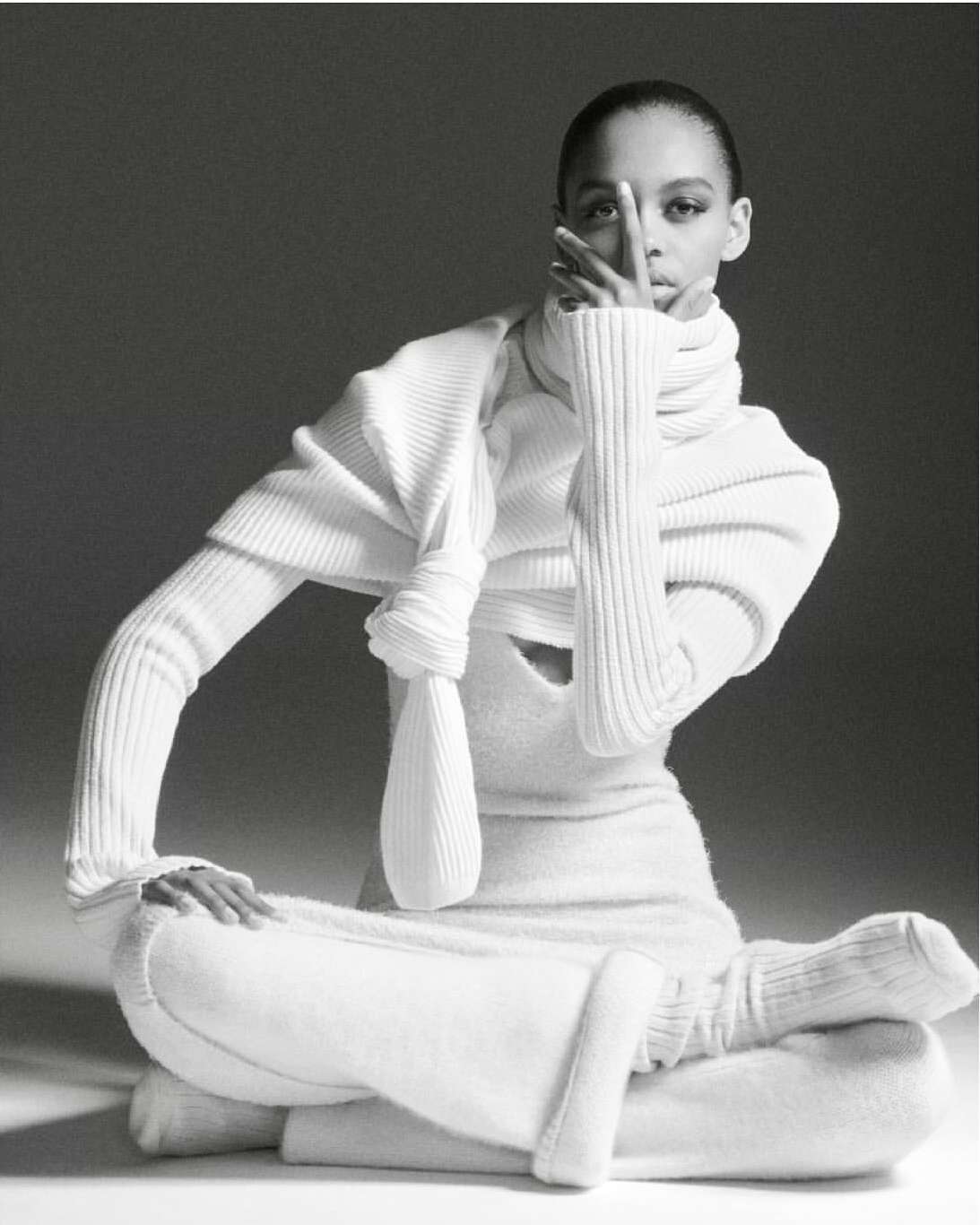 An adult female fashion model poses in a ribbed white turtleneck sweater and ribbed pants sitting cross-legged on a white surface.