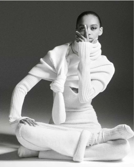An adult female fashion model poses in a ribbed white turtleneck sweater and ribbed pants sitting cross-legged on a white surface.