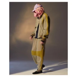Model wearing a light olive matching jacket and trousers with a pink patterned head covering and baseball cap.