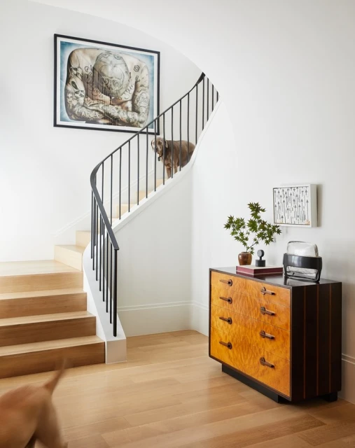 Hallway interior featuring a wooden staircase, a black metal banister, a light wood dresser, a framed art print, and a dog.