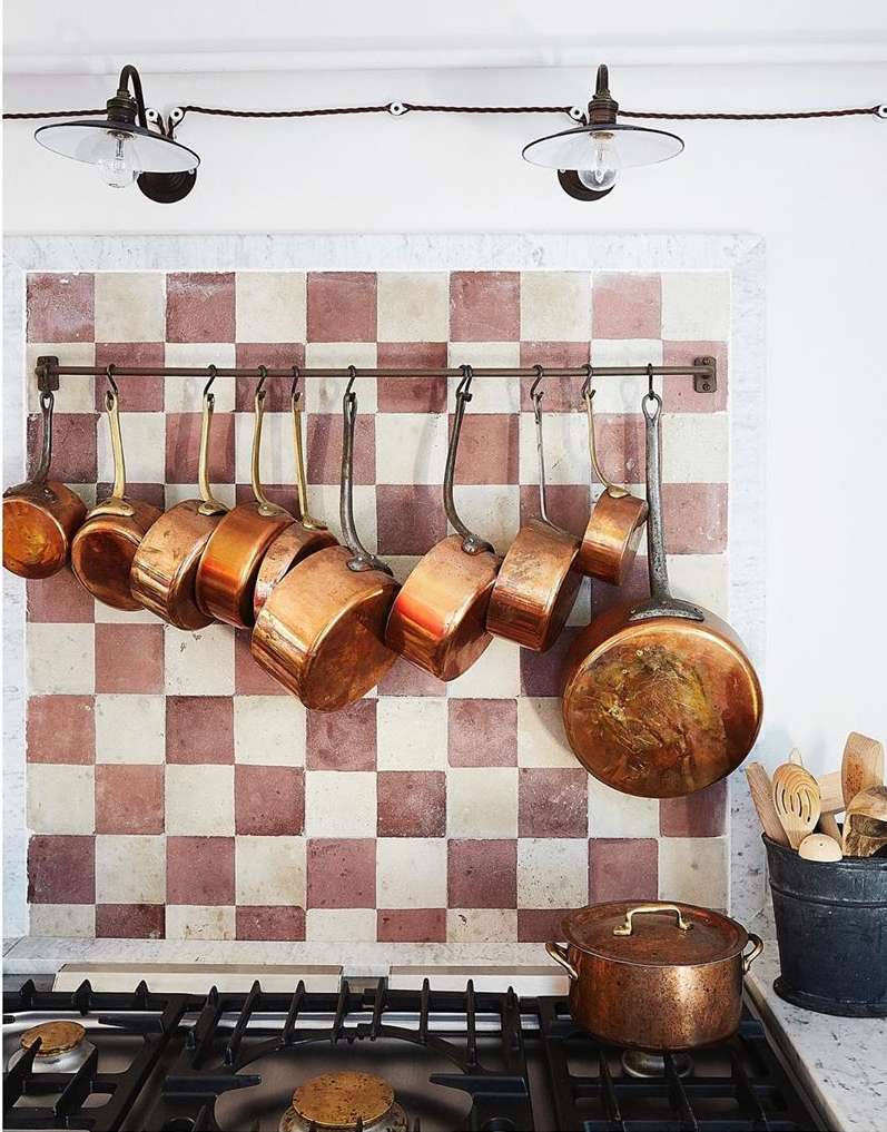 Kitchen interior with copper cookware hanging on a tile backsplash above a gas range, lit by overhead lamps, creating a warm, inviting space.