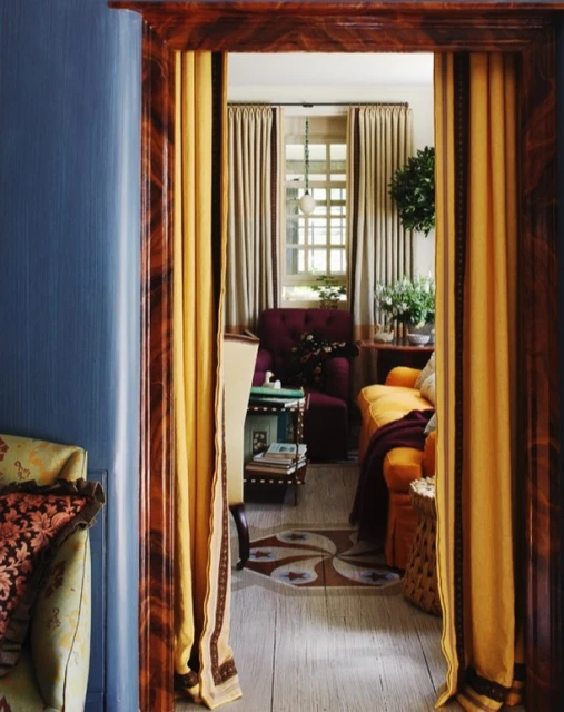 Ornate doorway frames living room scene with yellow curtains, plum and yellow seating, decorative area rug, and natural lighting.