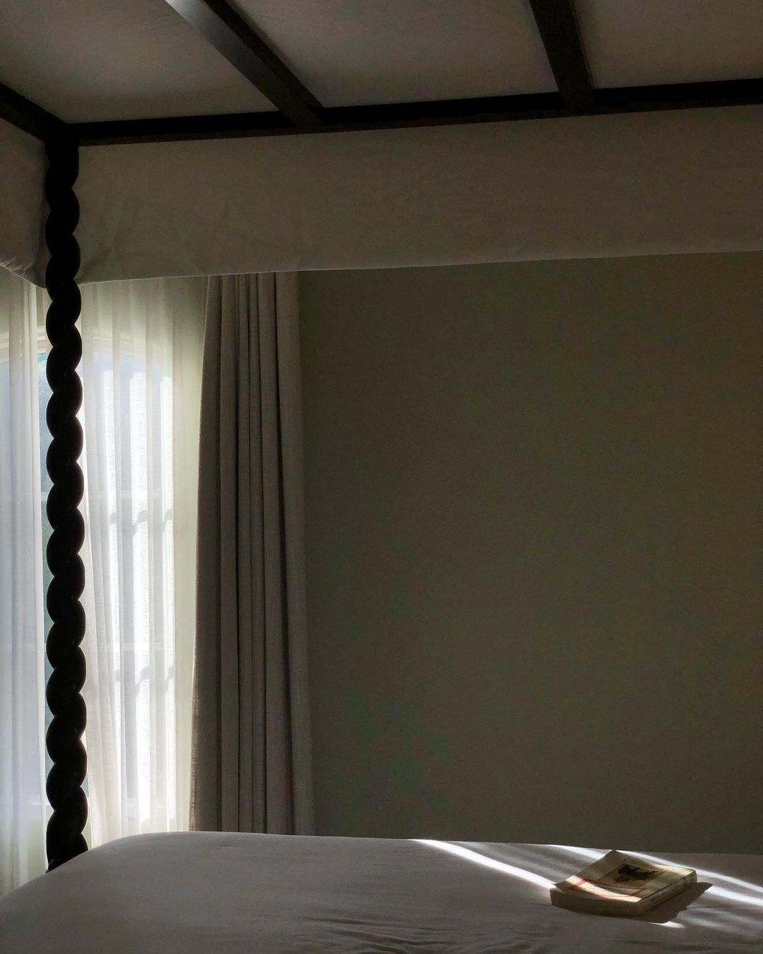 Four poster bed with white bedding, visible window, natural light, and a book on the bed in a bedroom interior.