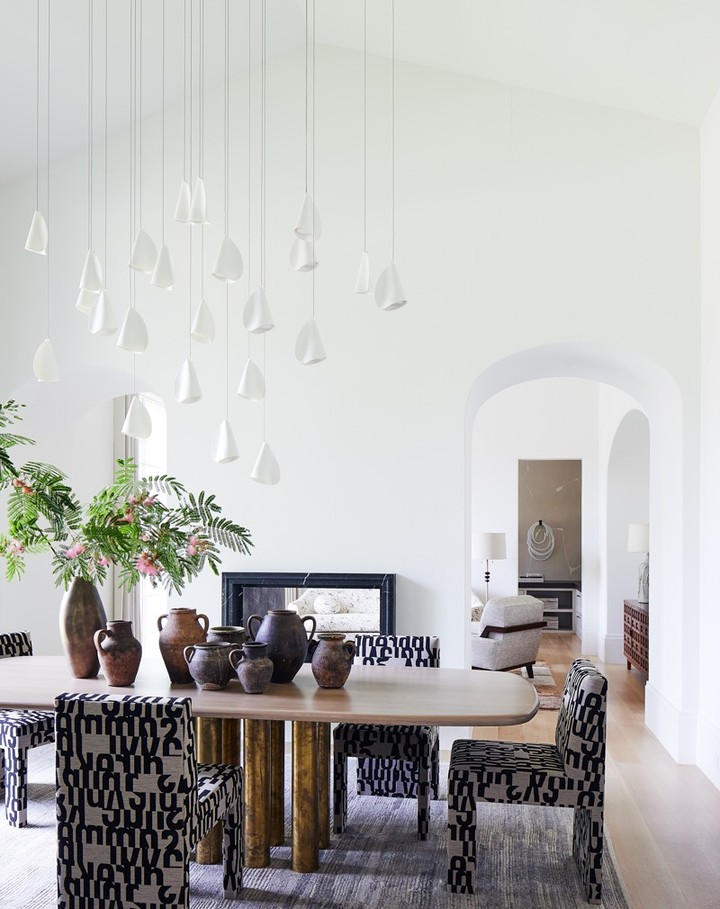 Dining room interior with a light wood table, patterned chairs, decorative vases, and unique pendant lighting against white walls.