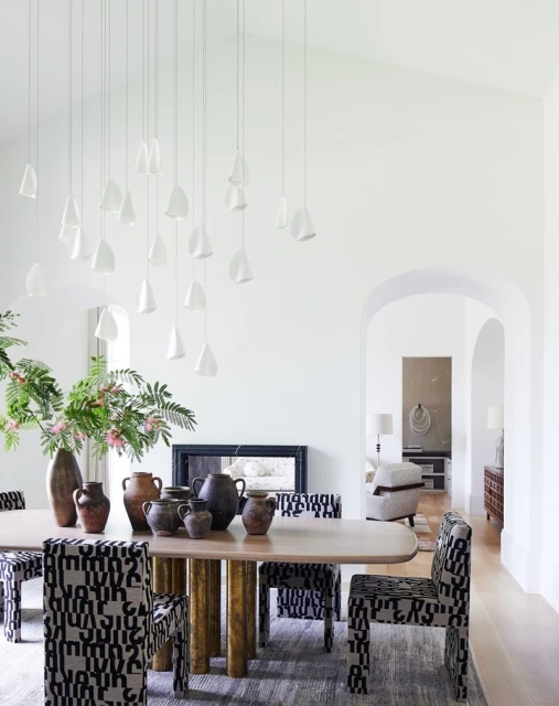Dining room interior with a light wood table, patterned chairs, decorative vases, and unique pendant lighting against white walls.