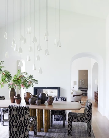 Dining room interior with a light wood table, patterned chairs, decorative vases, and unique pendant lighting against white walls.