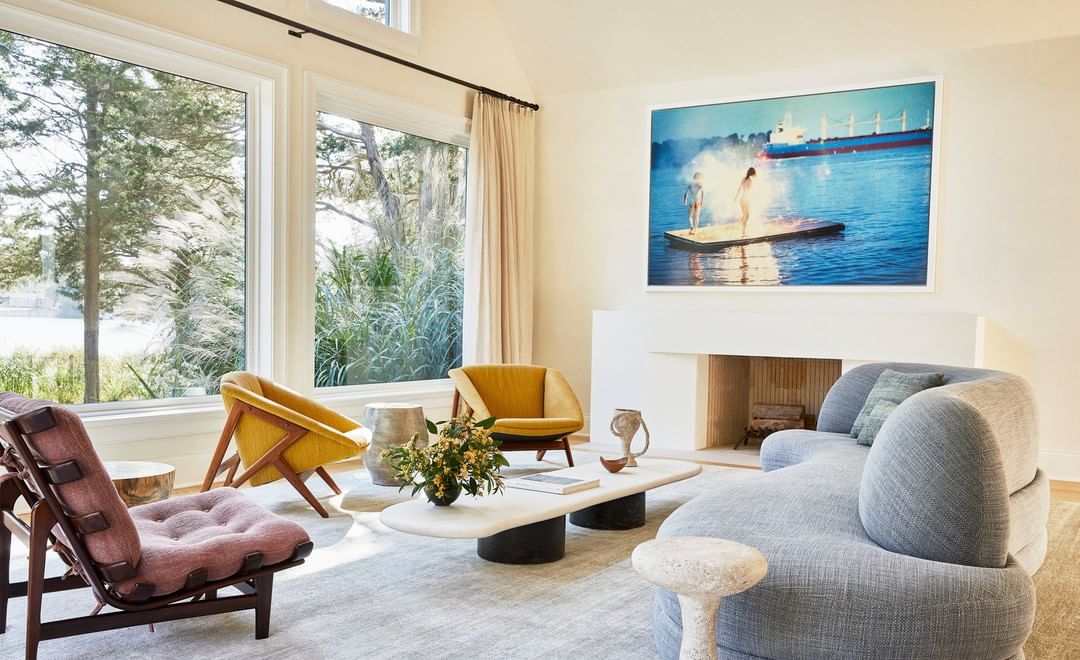 Living room interior featuring a modern gray sofa, chairs, a unique coffee table, and a fireplace, with light from the large windows.