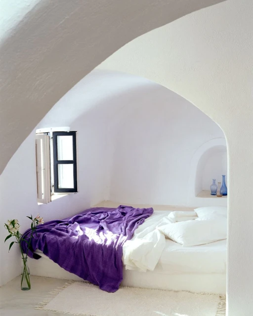 White arched bedroom with purple blanket, white linens, flowers, and a window with light coming in. Minimalist indoor architectural interior.