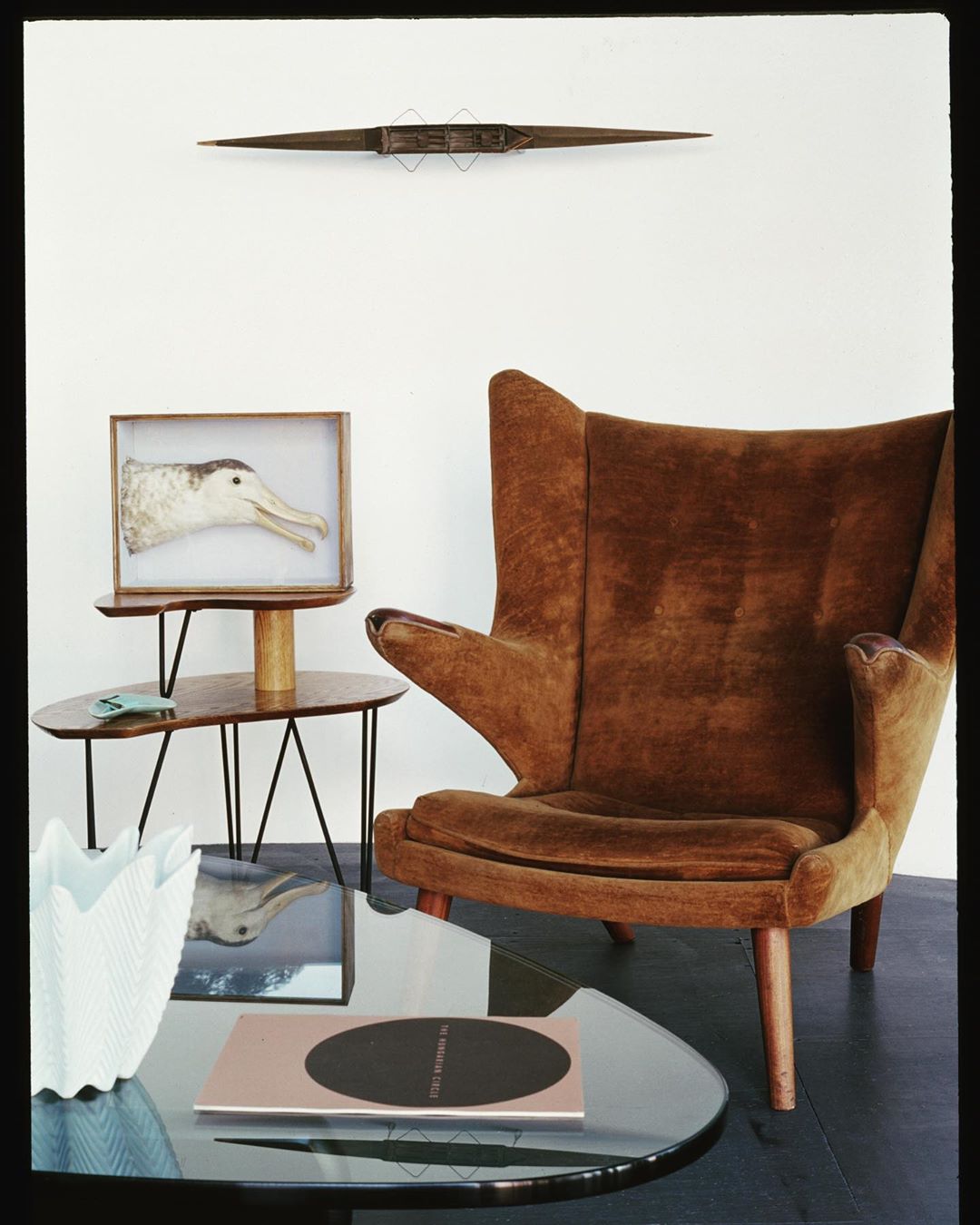 Interior with brown velvet armchair, modernist tables, and white walls in a neutral color scheme. Art objects adorn the scene.