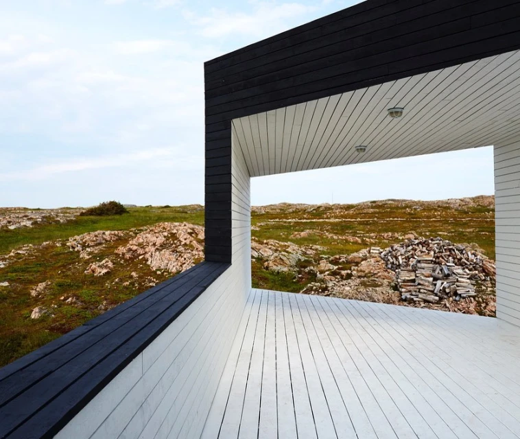 Architectural framing of a modern house with black and white wood, framing a field of rocks and grass under a blue sky.