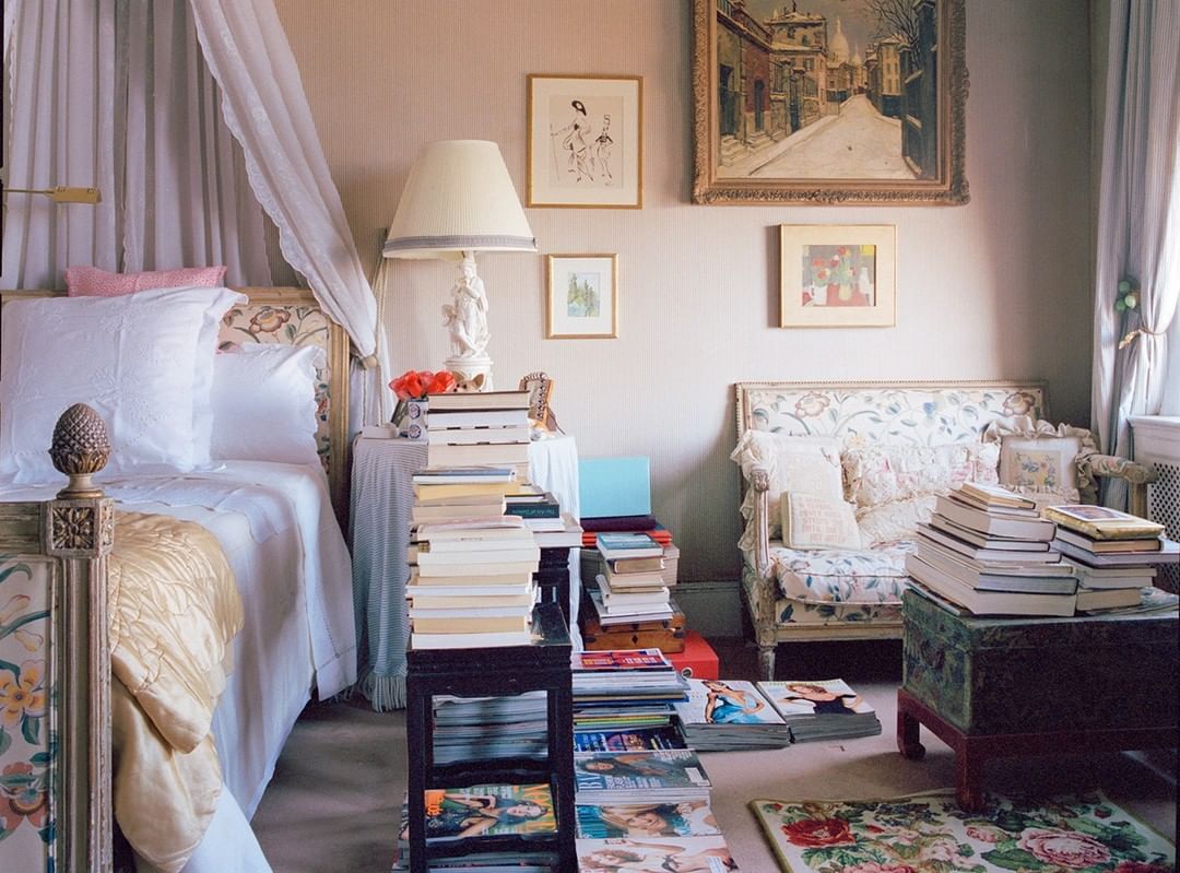Bedroom with a floral sofa, a bed draped in white fabric, art on the walls, and stacks of books and magazines throughout the room.