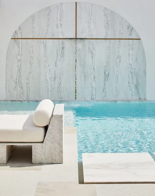 Outdoor scene featuring a daybed beside a turquoise pool, complemented by marble steps and a textured white wall with arched details.