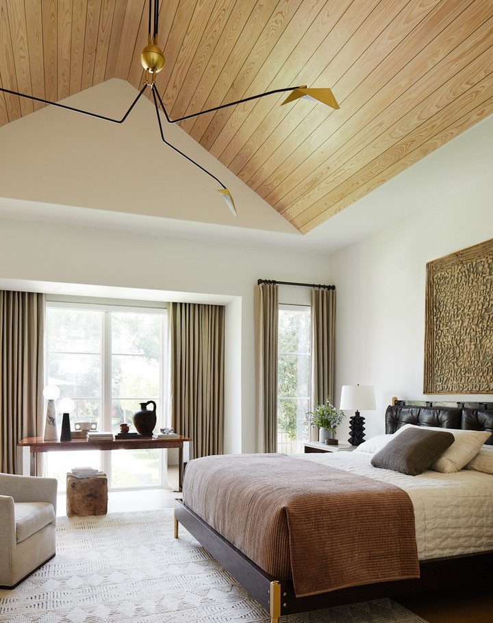 Bedroom interior with a bed, desk with decorative items, wooden floors and ceiling, large windows, and modern black light fixture.