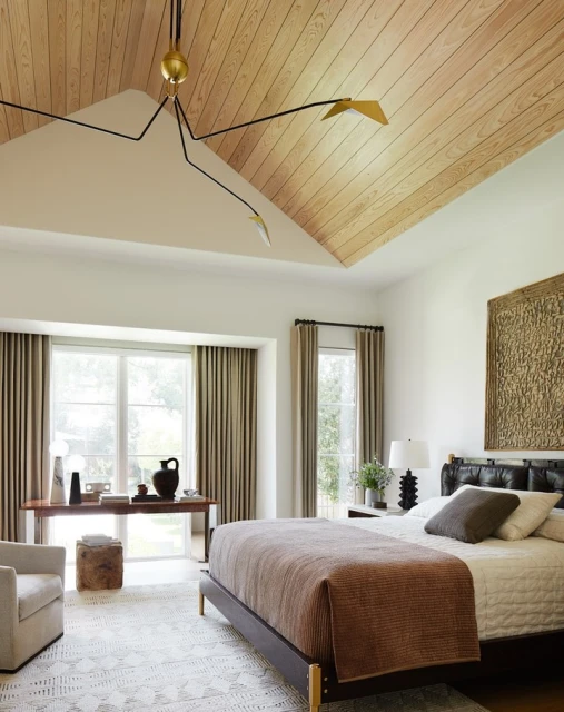 Bedroom interior with a bed, desk with decorative items, wooden floors and ceiling, large windows, and modern black light fixture.