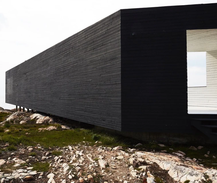 Exterior of a modern black wooden building on a rocky, grassy landscape under a bright sky. The building has a minimalist design.