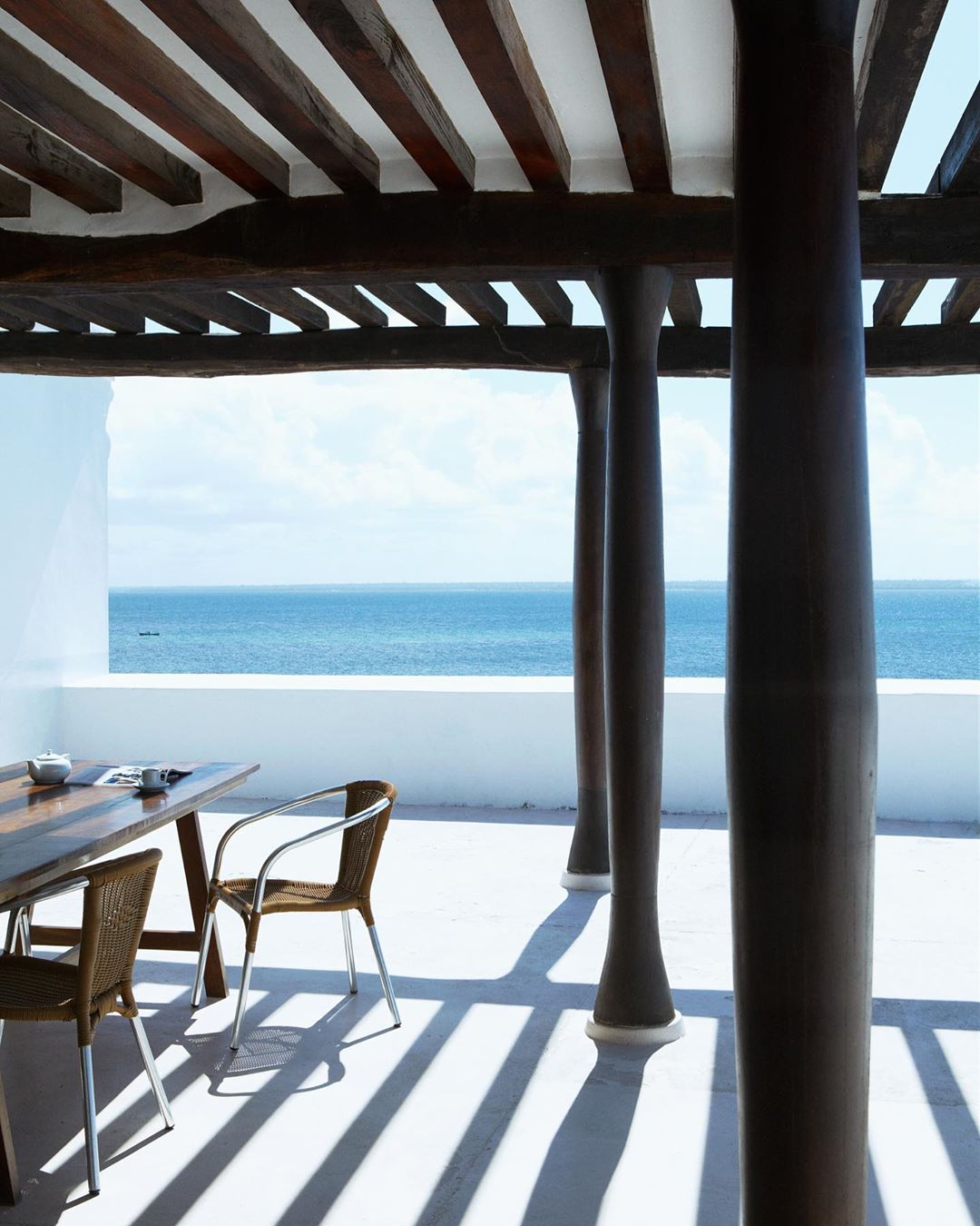 A patio with chairs and a table under a wood-beamed roof overlooks a tranquil ocean view on a bright day, showcasing architectural design.