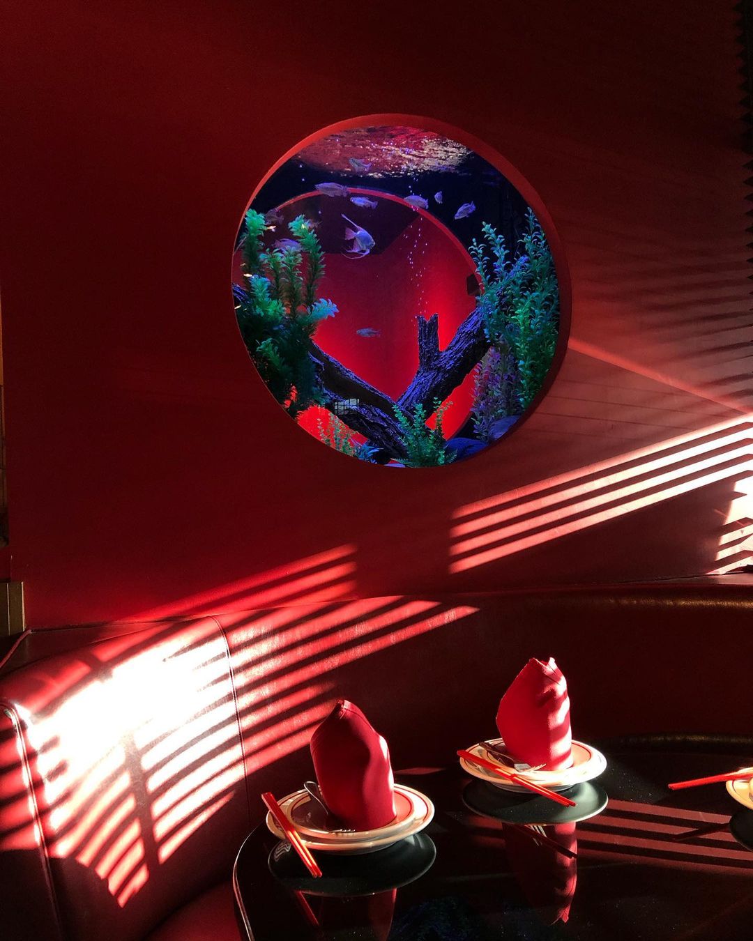 Fish swim in a round aquarium mounted on a red wall. Below, a table is set with red napkins and silverware, illuminated by light through blinds.