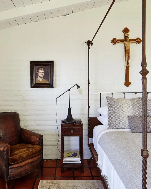 Bedroom interior featuring a wrought-iron bed, dark wood nightstand with a lamp, vintage-style artwork, and a brown leather armchair.