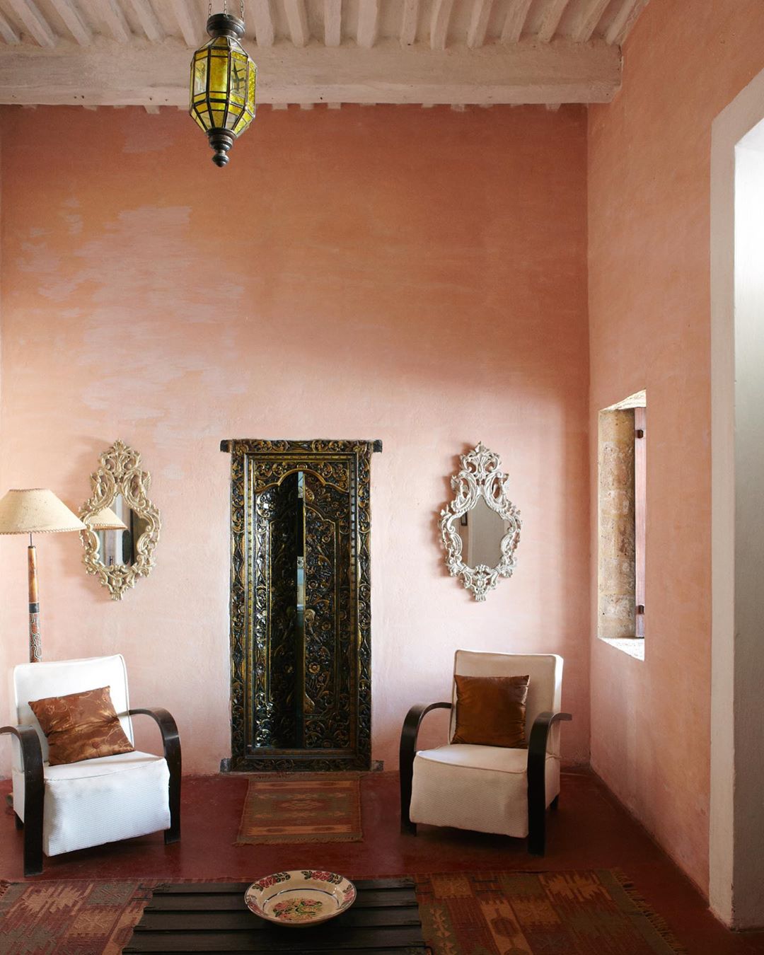 Interior of a room with pink walls, featuring two armchairs, an ornate black door, decorative mirrors, and a hanging lantern.