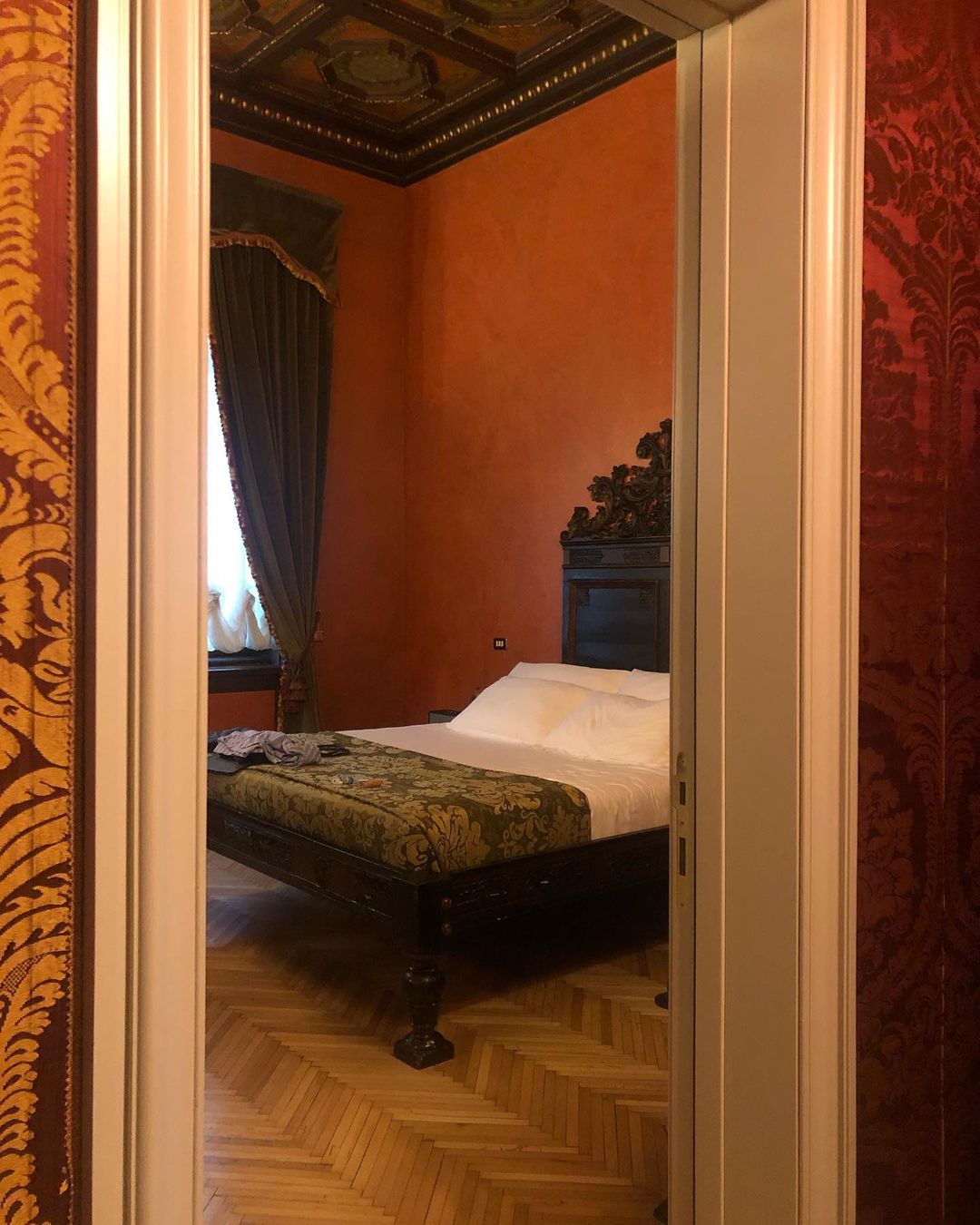 View of a bedroom with a carved wooden headboard and a green patterned bench at the foot of the bed, seen through an open doorway.