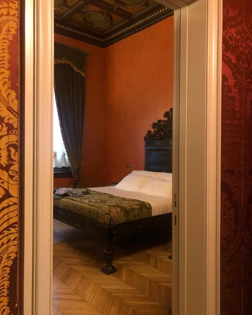 View of a bedroom with a carved wooden headboard and a green patterned bench at the foot of the bed, seen through an open doorway.