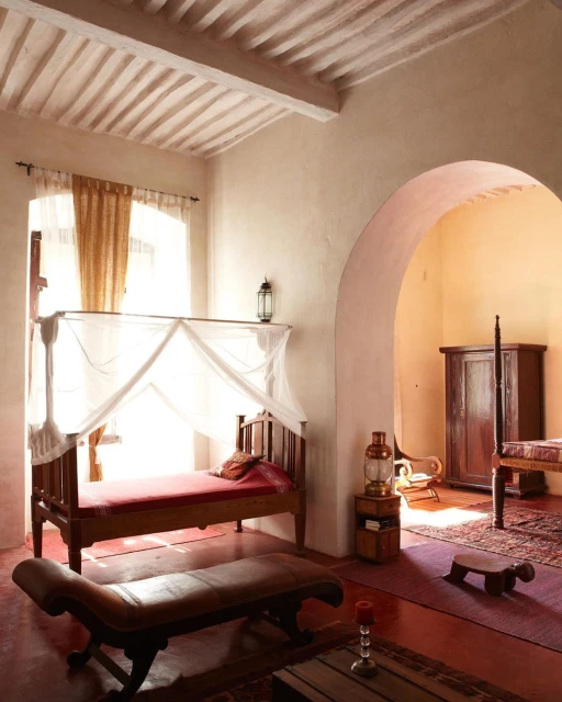 Indoor bedroom with a four-poster bed with mosquito net, lounger, decorative lighting, and arched doorway showcasing additional beds.