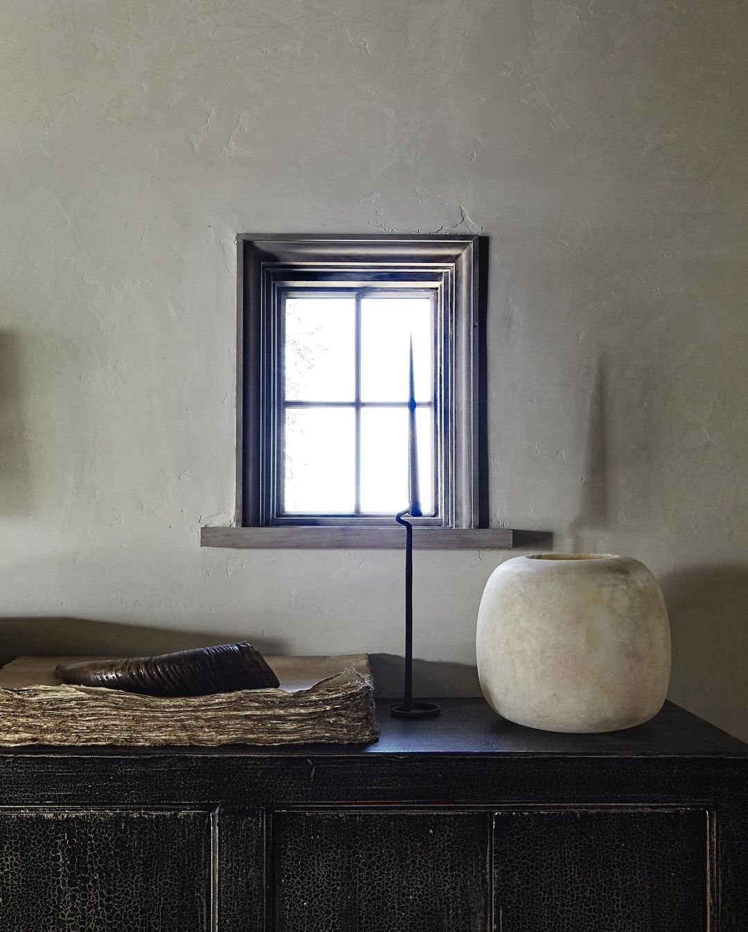 Interior still life featuring a vase, book, and candleholder on a black cabinet against a neutral wall with a window.