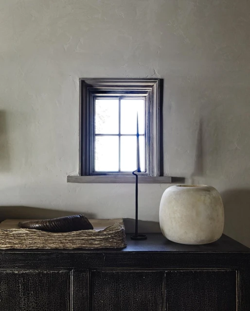 Interior still life featuring a vase, book, and candleholder on a black cabinet against a neutral wall with a window.