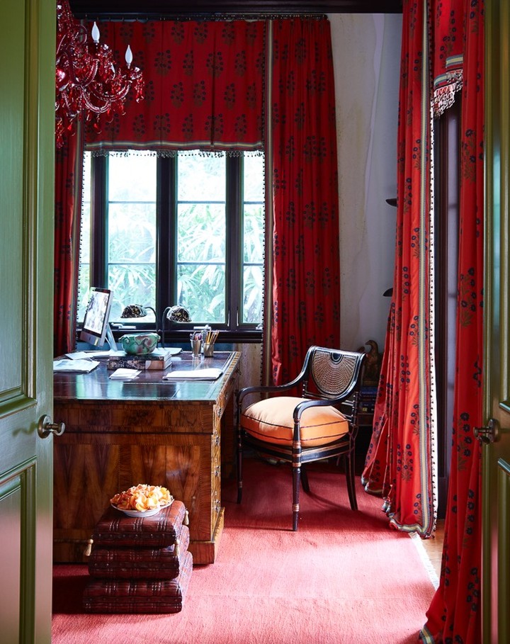 A wood desk with computer, books, and chair are positioned in a room with red patterned drapes, carpet, and a red chandelier.