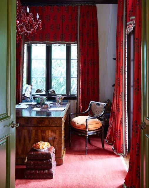 A wood desk with computer, books, and chair are positioned in a room with red patterned drapes, carpet, and a red chandelier.