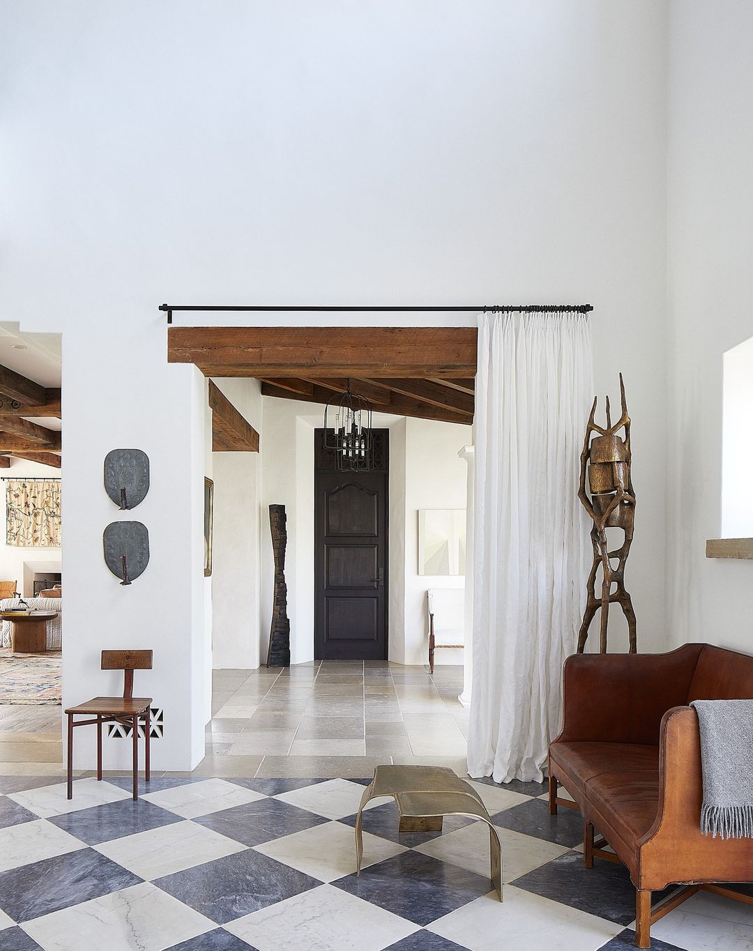 Interior view of a living room with a checkered tile floor, a brown sofa, a wooden chair, and modern sculptures in a bright, naturally lit space.