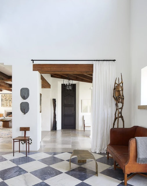Interior view of a living room with a checkered tile floor, a brown sofa, a wooden chair, and modern sculptures in a bright, naturally lit space.
