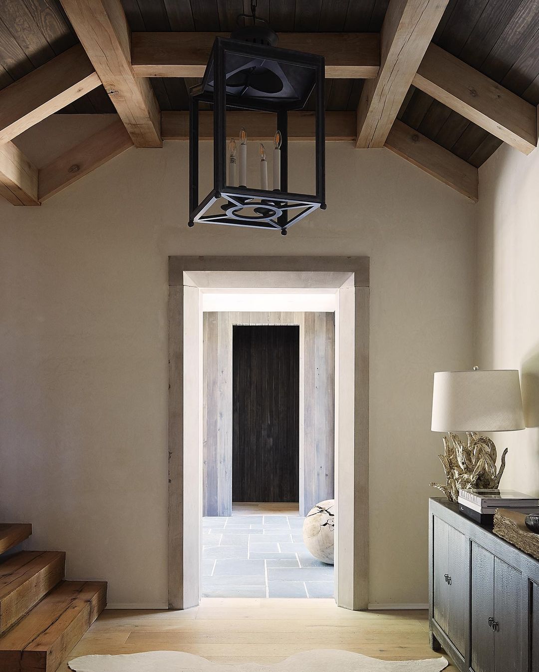 Interior view of an entryway with light walls and ceiling, wooden beams, a modern black light fixture, and a dark wood credenza.