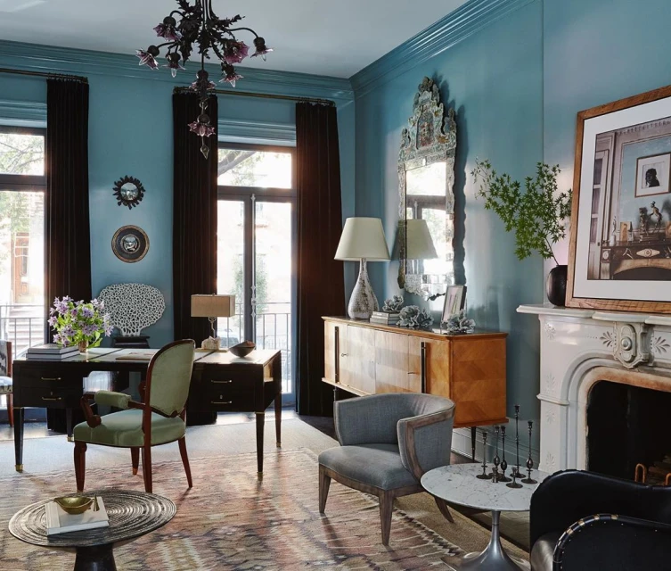 Living room interior showcasing a desk setup, grey upholstered chair, fireplace, mirror, and a patterned area rug under soft, diffused light.