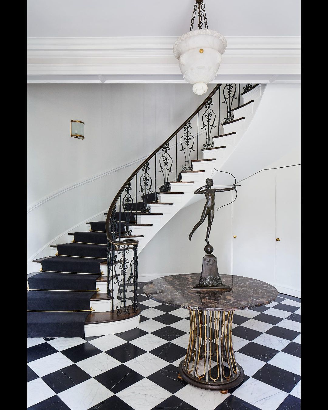 Interior view of a home's entryway featuring a staircase with black carpet, statue on a table, chandelier, and black-and-white checkered floor.