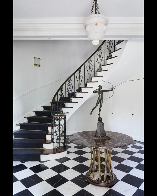 Interior view of a home's entryway featuring a staircase with black carpet, statue on a table, chandelier, and black-and-white checkered floor.