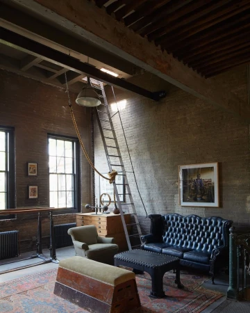 Interior of a vintage-style industrial space featuring a black leather sofa, armchair, exercise equipment, and exposed brick walls.