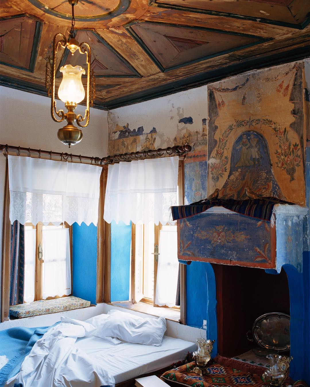 Bedroom interior featuring a bed with white linens, decorative blue walls, and a painted ceiling with a hanging lamp above the bed.