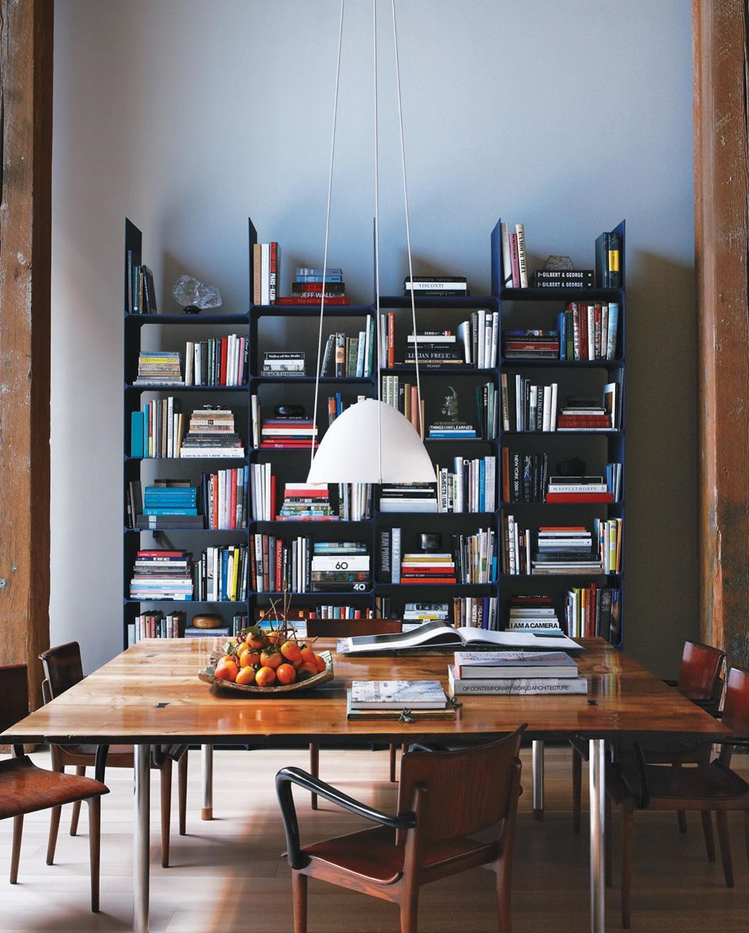 A dining room features a wooden table surrounded by chairs, illuminated by a modern pendant light, with a tall bookshelf in the background.