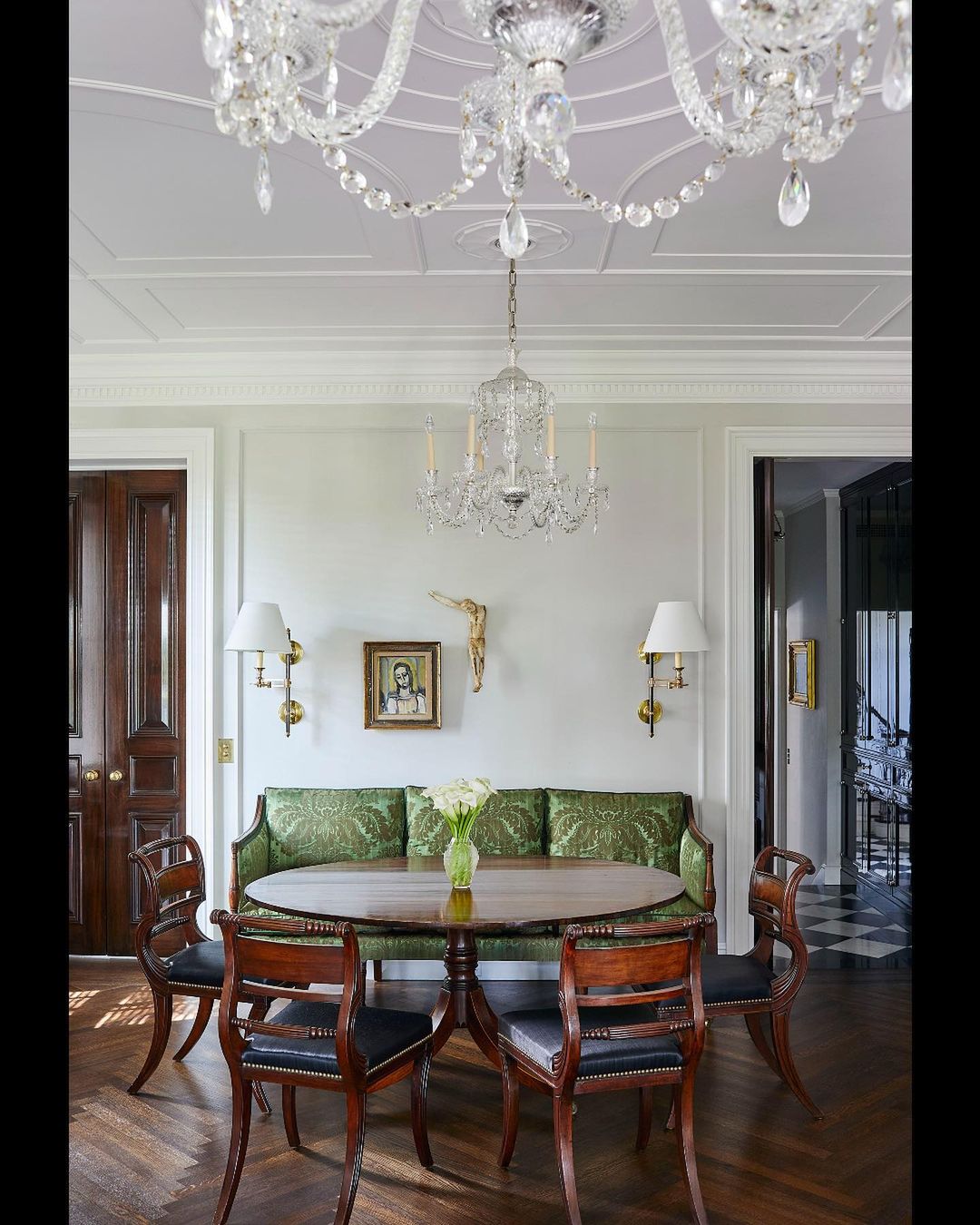 Interior view of a dining area with a round wood table, upholstered chairs, and crystal chandeliers providing soft, ambient lighting.
