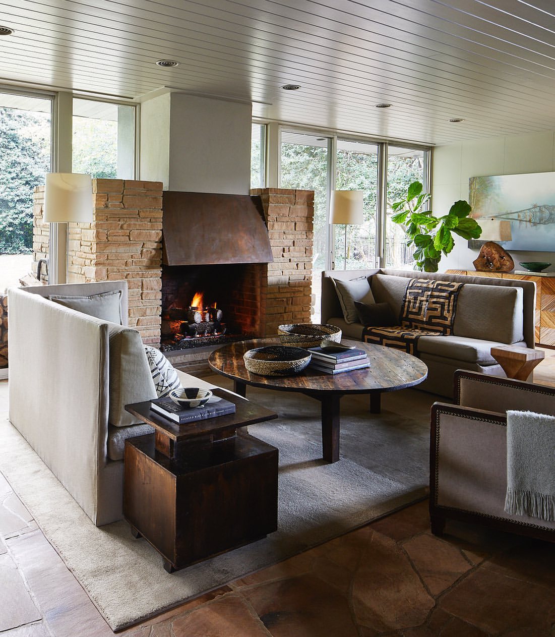 Living room interior featuring stone fireplace, neutral couches, wooden coffee table with books, and view of green trees through windows.