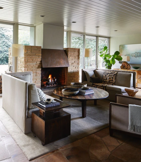 Living room interior featuring stone fireplace, neutral couches, wooden coffee table with books, and view of green trees through windows.