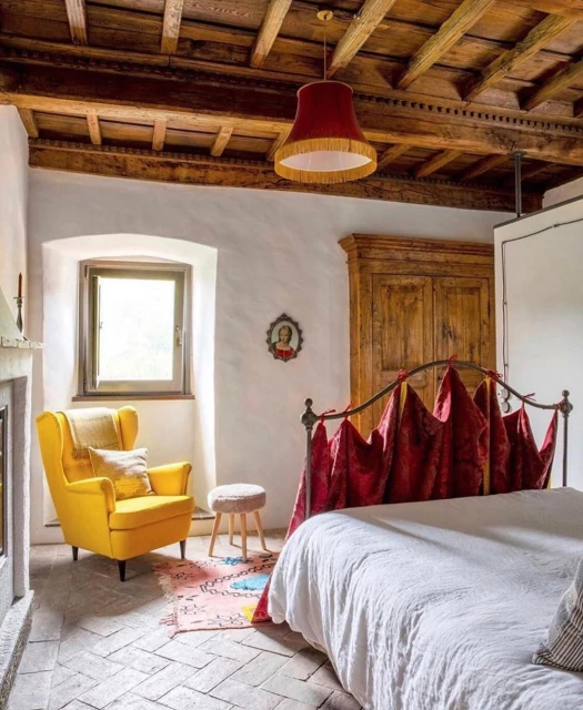 Cozy bedroom interior featuring a bed with red accents, a yellow armchair, exposed wood beam ceiling, and neutral-toned walls and floors.