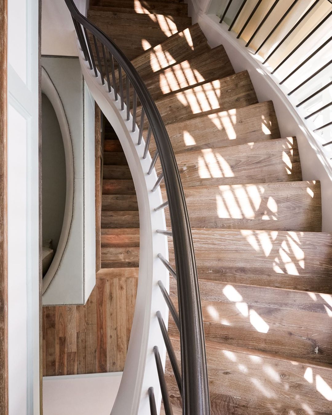 Interior view of a wooden staircase with natural light casting shadows, complemented by a white wall and banister with black metal accents.