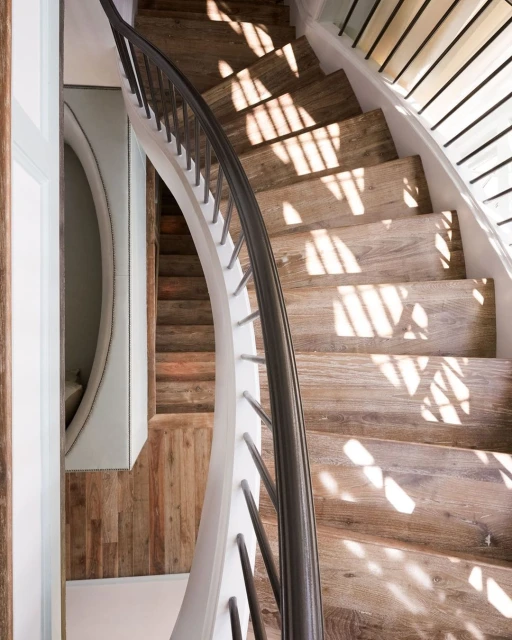 Interior view of a wooden staircase with natural light casting shadows, complemented by a white wall and banister with black metal accents.