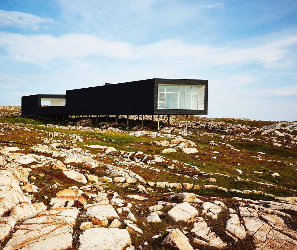 Modern black house with glass windows on stilts, set on rocky landscape with sparse vegetation, clear sky. Architecture, design.