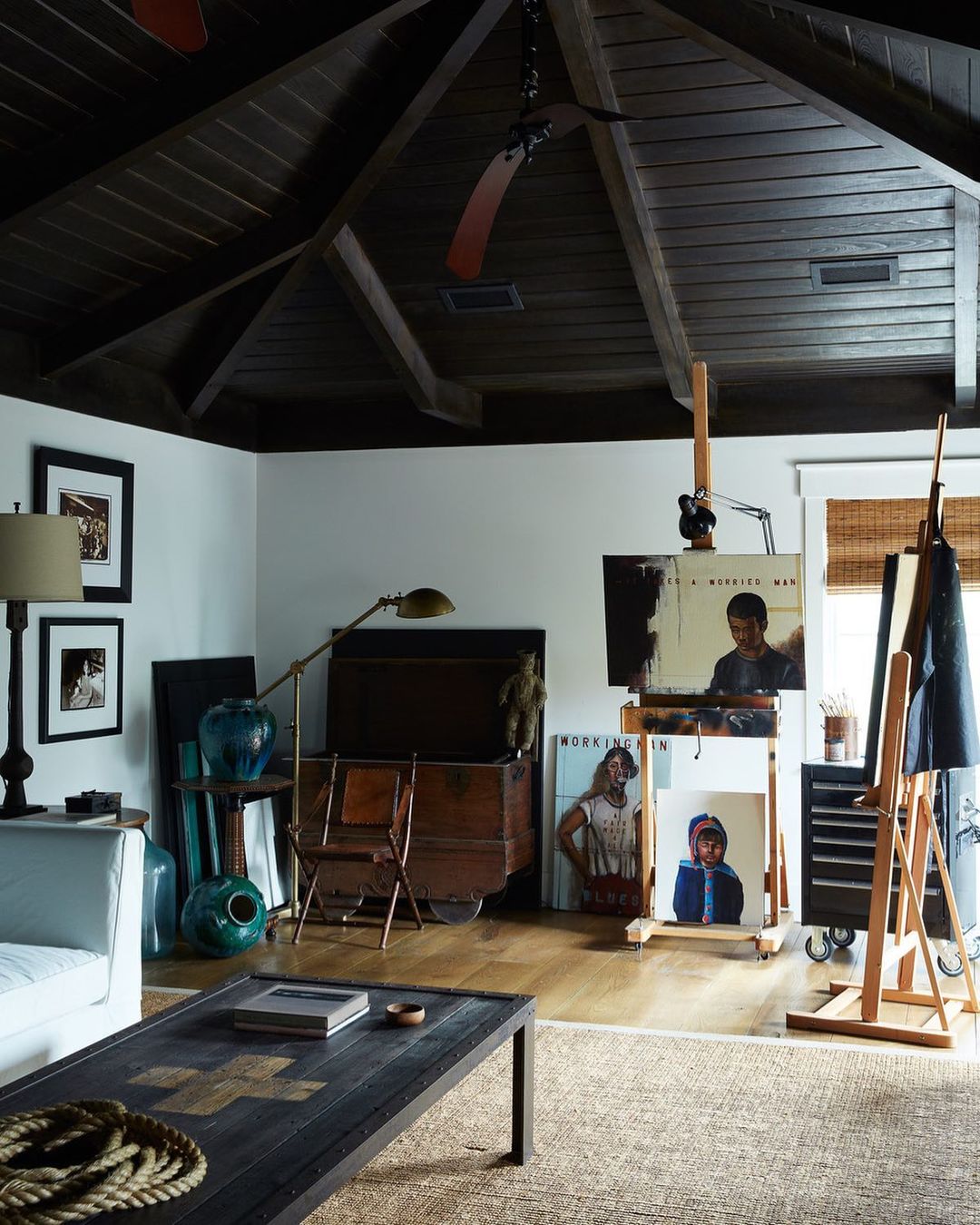 Living room interior featuring paintings on easels, a dark wood ceiling, neutral walls, and eclectic furniture in a well-lit space.