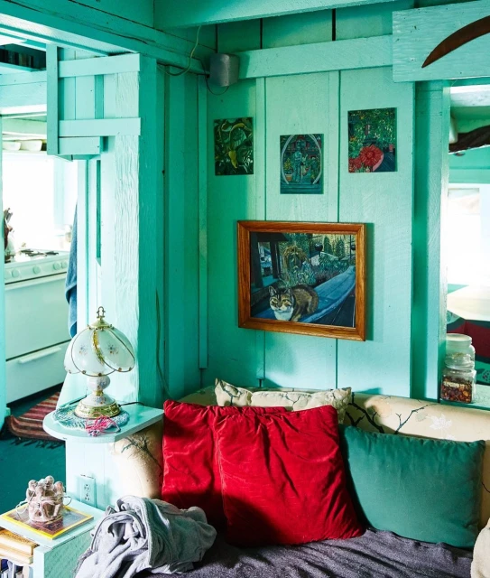 Interior view of a brightly colored aqua room featuring pillows, a lamp, paintings, and rustic decor with a cozy, lived-in aesthetic.